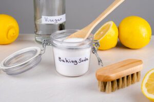 A clear jar of baking soda with a wooden spoon in it, surrounded by a bottle of vinegar, lemons, and a cleaning brush.